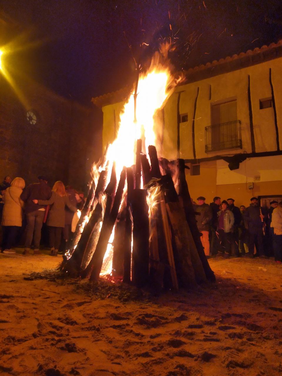 Apilamiento de troncos ardiendo en torno a un pino situado en el centro de la plazuela del Doncel. La gente se agolpa en torno a la hoguera, con el exterior de la iglesia de San Vicente como fondo